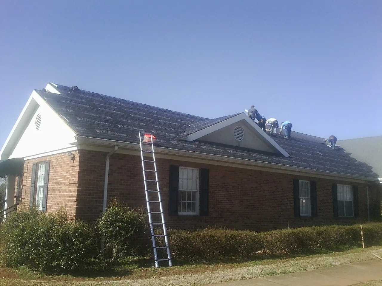 Crew tearing shingles off a brick ranch roof with a ladder set — Emergency Roof Repair work by our crew in Brewerton
