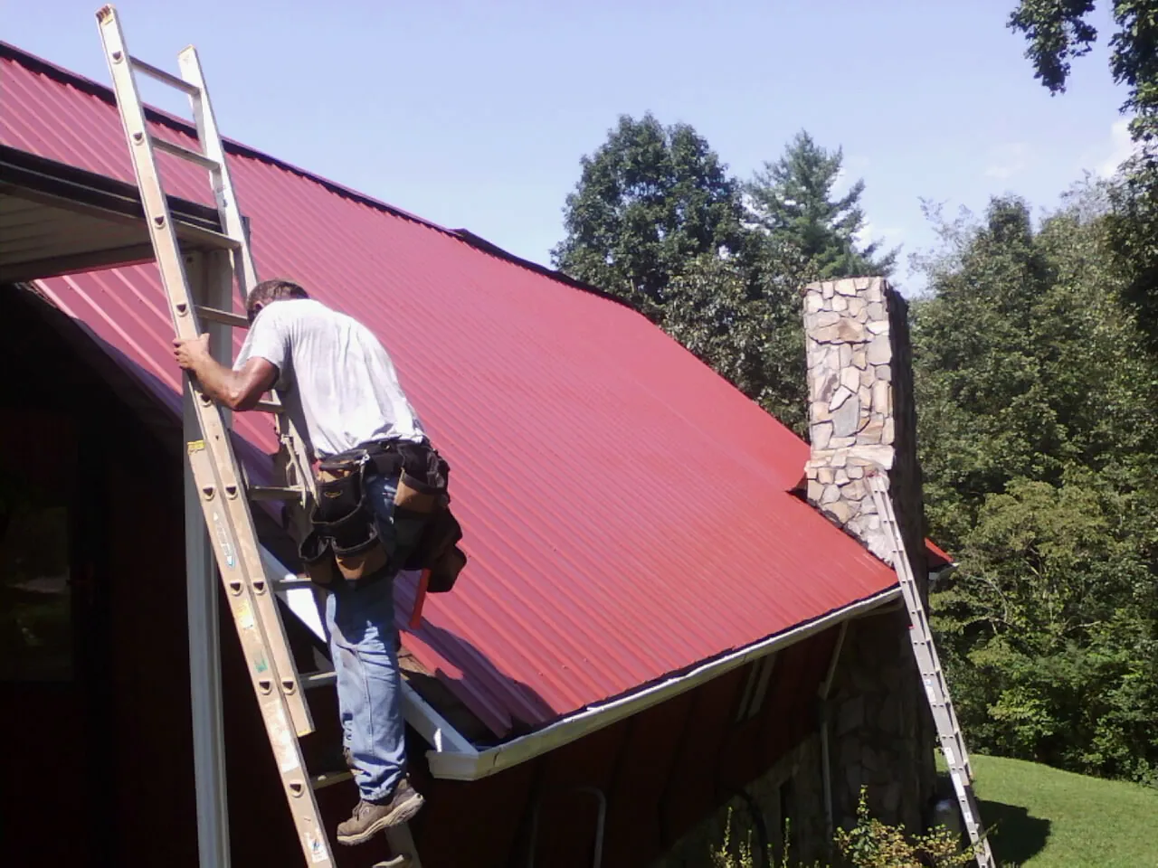 Roofer on a ladder beside a red metal roof and stone chimney — Soffit & Fascia Repair by Roofing Brothers in Brewerton